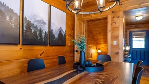 Un interior de madera con una larga mesa de comedor, sillas, un jarrón con follaje verde y cuadros en la pared.