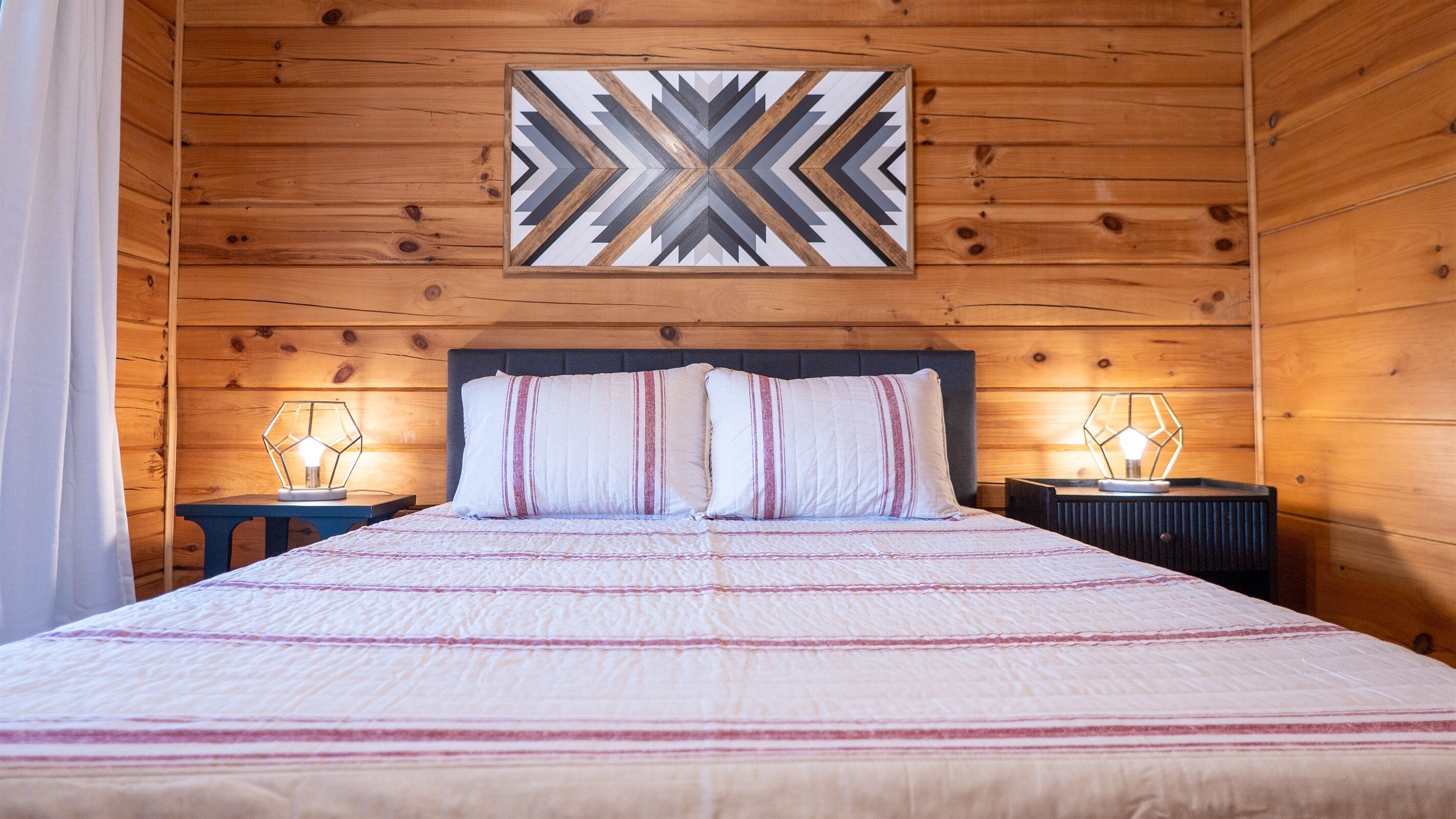 A wooden cabin-style bedroom with a large bed, two bedside tables, and a geometric wall art piece.