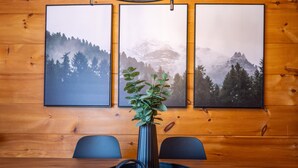 Un interior de madera con una mesa de comedor, dos sillas, un jarrón con una planta y tres cuadros en la pared.