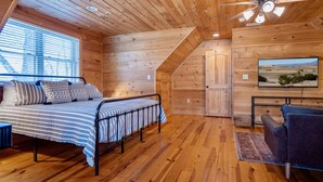 Una habitación acogedora tipo cabaña de madera, con una cama, una silla y un televisor que muestra un paisaje desértico.
