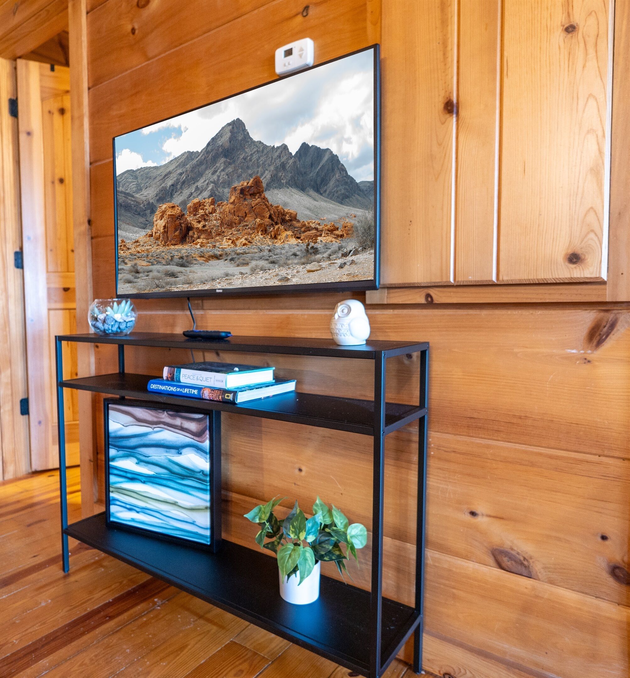 Un interno in legno con una televisione a parete che mostra un paesaggio montano, un mobile intrattenimento nero con libri e una pianta, e un piccolo oggetto decorativo bianco sopra il mobile.