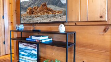 Un interno in legno con una televisione a parete che mostra un paesaggio montano, un mobile intrattenimento nero con libri e una pianta, e un piccolo oggetto decorativo bianco sopra il mobile.