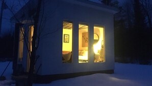 A small, single-story cabin with a visible bedroom and a lit interior, surrounded by snow. - Glamping hut with WiFi in Ripton, biking, hiking, skiing (Ripton)