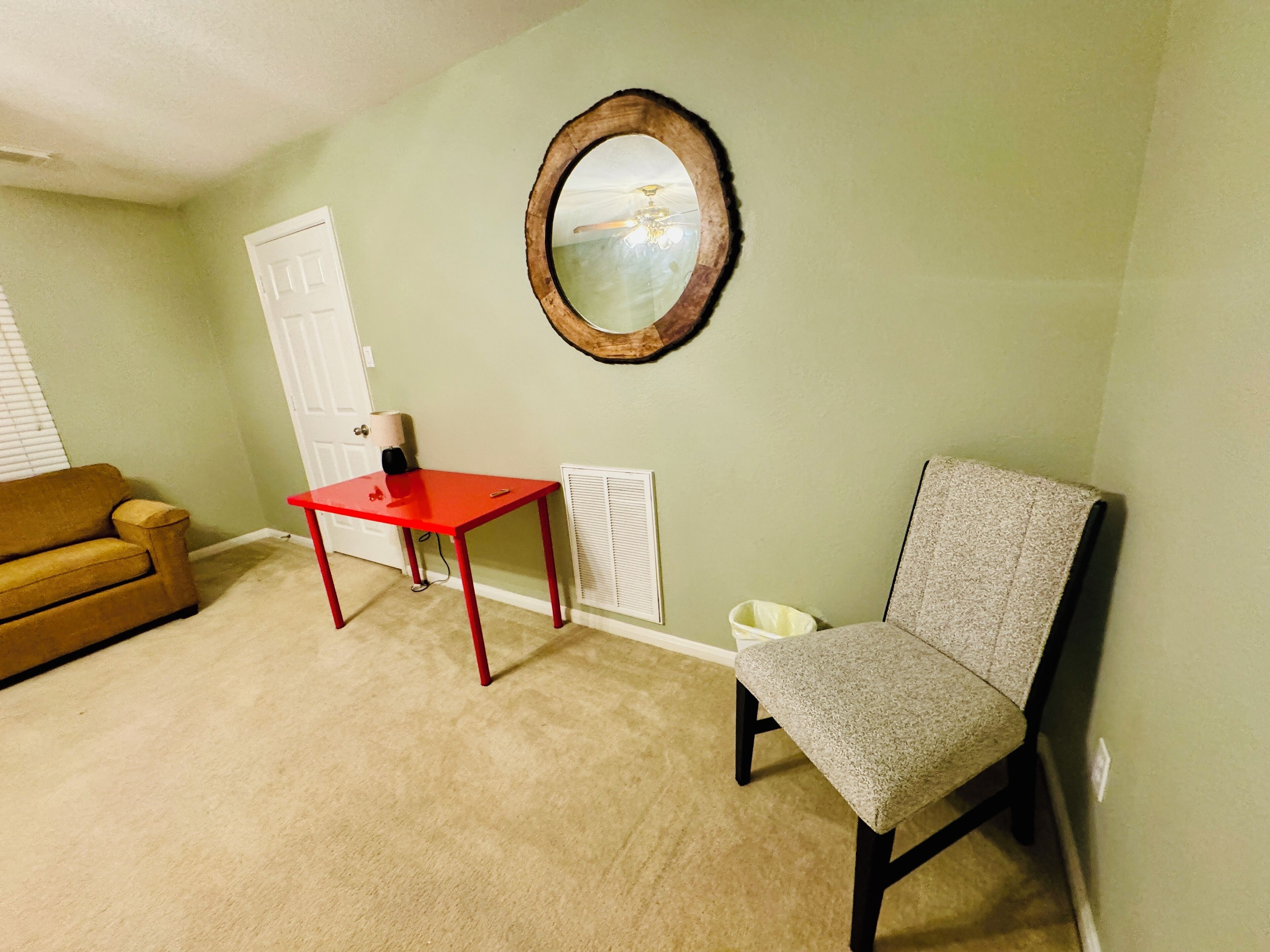 Ein Zimmer mit beigeem Teppich, einem roten Tisch, einem grauen Stuhl, einer weißen Tür und einem runden Spiegel an der Wand.