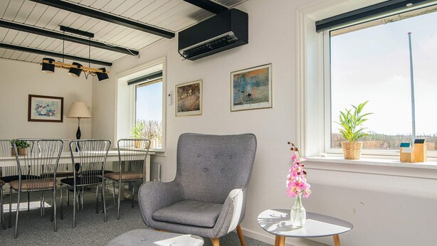 A modern room with a grey armchair, a small round table with a vase of flowers, a dining area with metal chairs, and a large window with a view. - 5 Person Holiday Home in Snedsted (Snedsted)