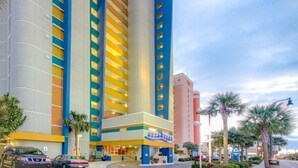 Un hotel de varios pisos con una fachada llamativa en azul y amarillo, una entrada central y palmeras en primer plano.
