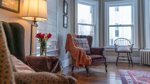 A cozy living room with a brown armchair, a wooden chair, and a lamp. - Historic 4-bedroom house one block from Saratoga Racetrack (Saratoga Springs)