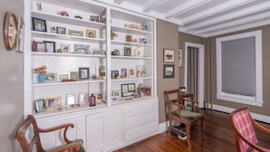 A room with a white built-in bookshelf filled with various framed photos and decorative items, wooden chairs, and a large window with blinds. - Historic 4-bedroom house one block from Saratoga Racetrack (Saratoga Springs)