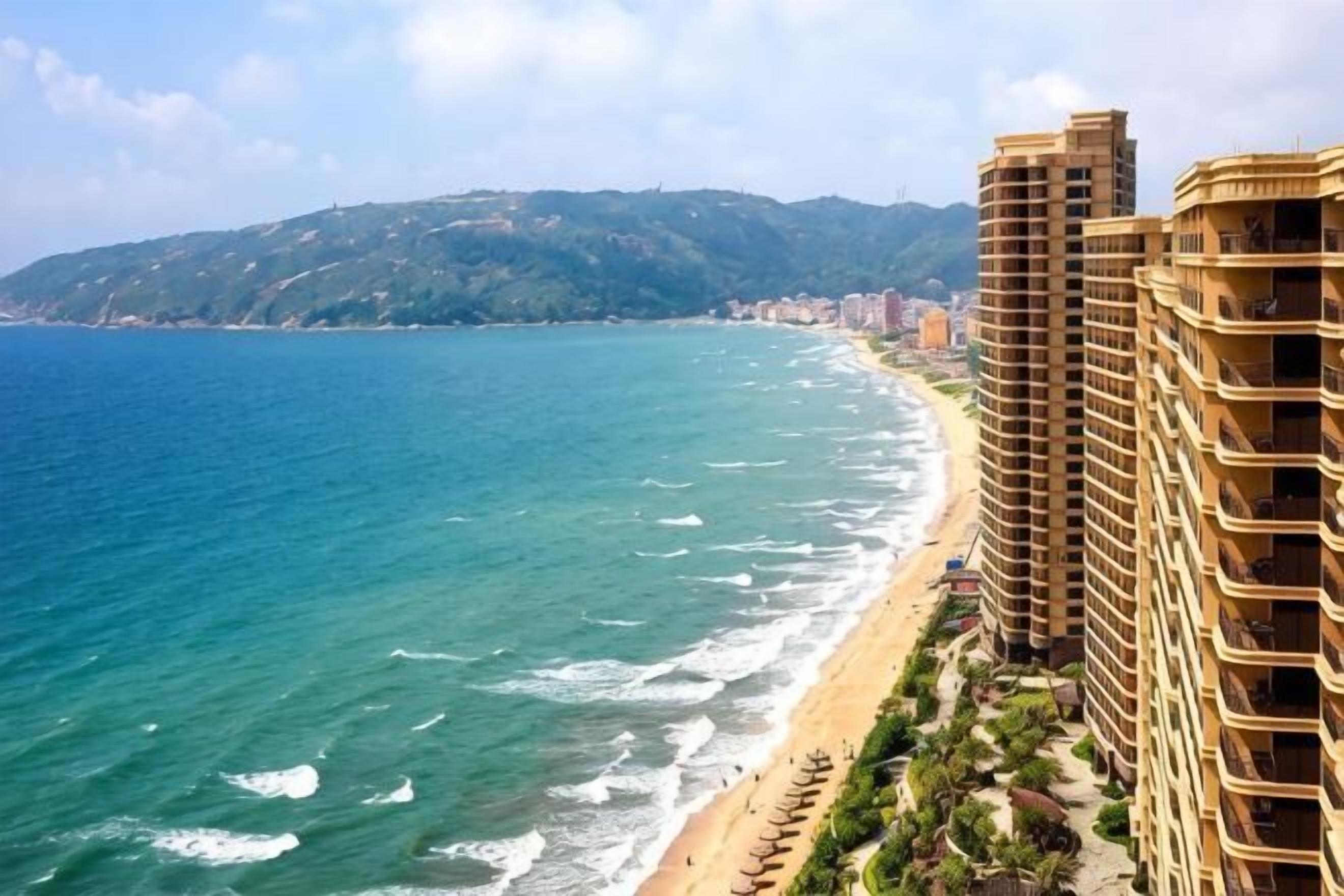 Un hotel frente al mar con vista a la playa, edificios altos y un paisaje montañoso al fondo.