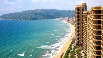 Un hotel frente al mar con vista a la playa, edificios altos y un paisaje montañoso al fondo.
