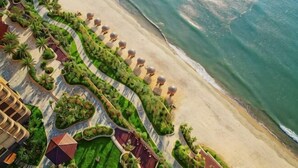 Un hôtel situé en bord de mer, avec un long sentier sinueux bordé d’arbres et de parasols, qui mène à une plage de sable.