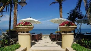 Un chemin mène à un espace extérieur aménagé avec des parasols et des jardinières fleuries, offrant une vue sur la mer.