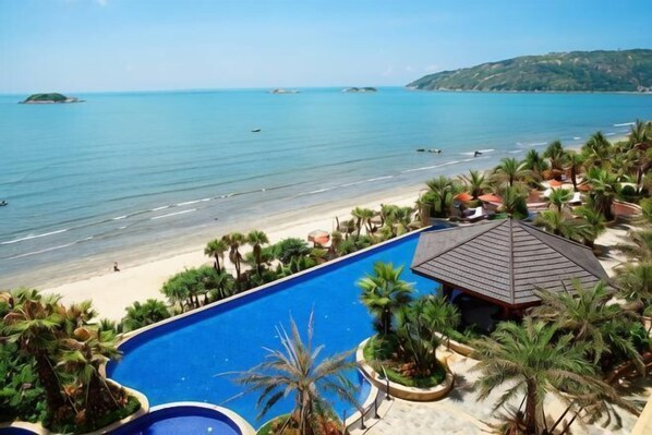 A pool with a view of the beach and ocean, surrounded by palm trees and a small building.