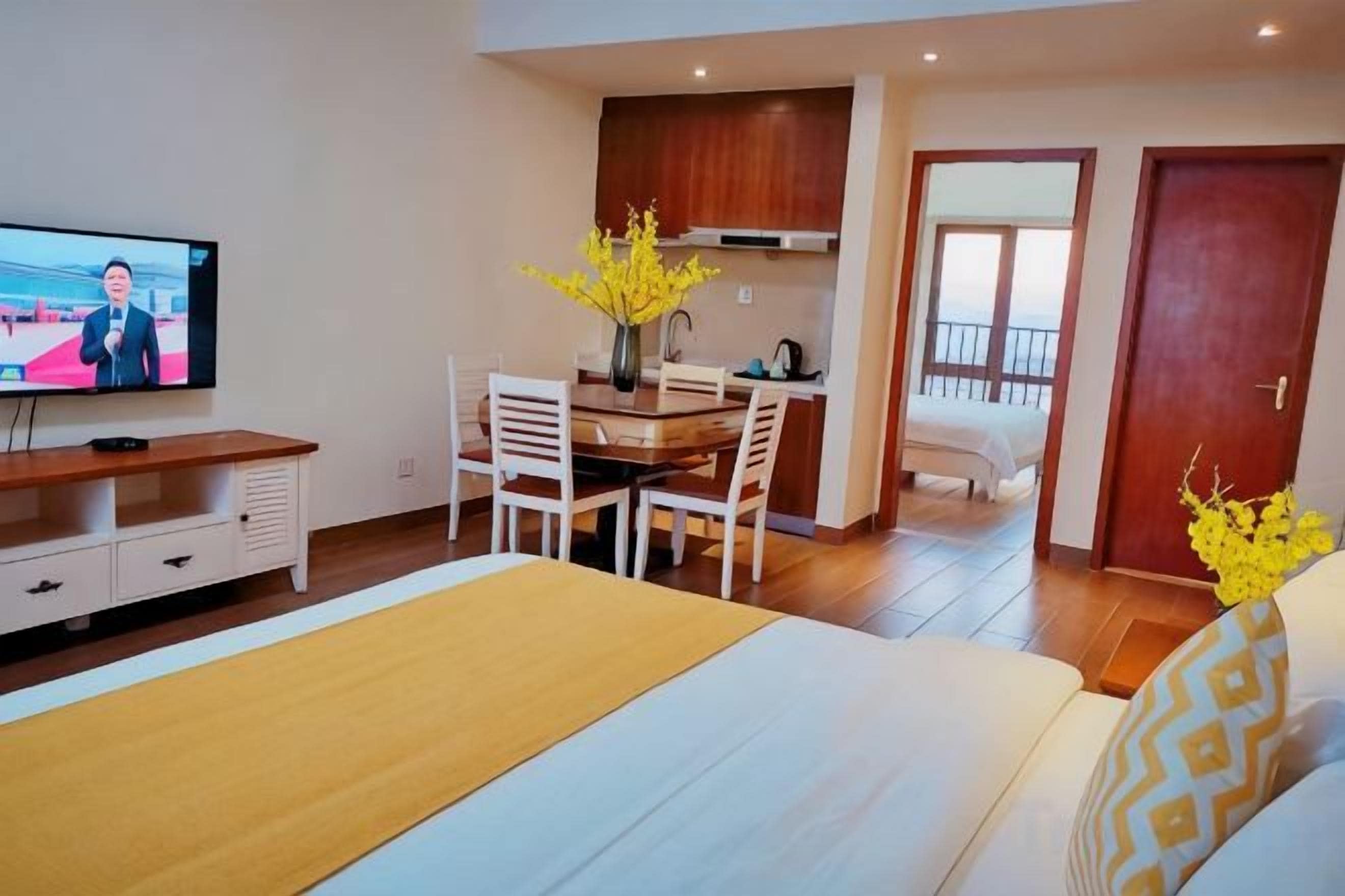 A hotel room with a bed, a TV, a dining table with chairs, a kitchen area, and a balcony.