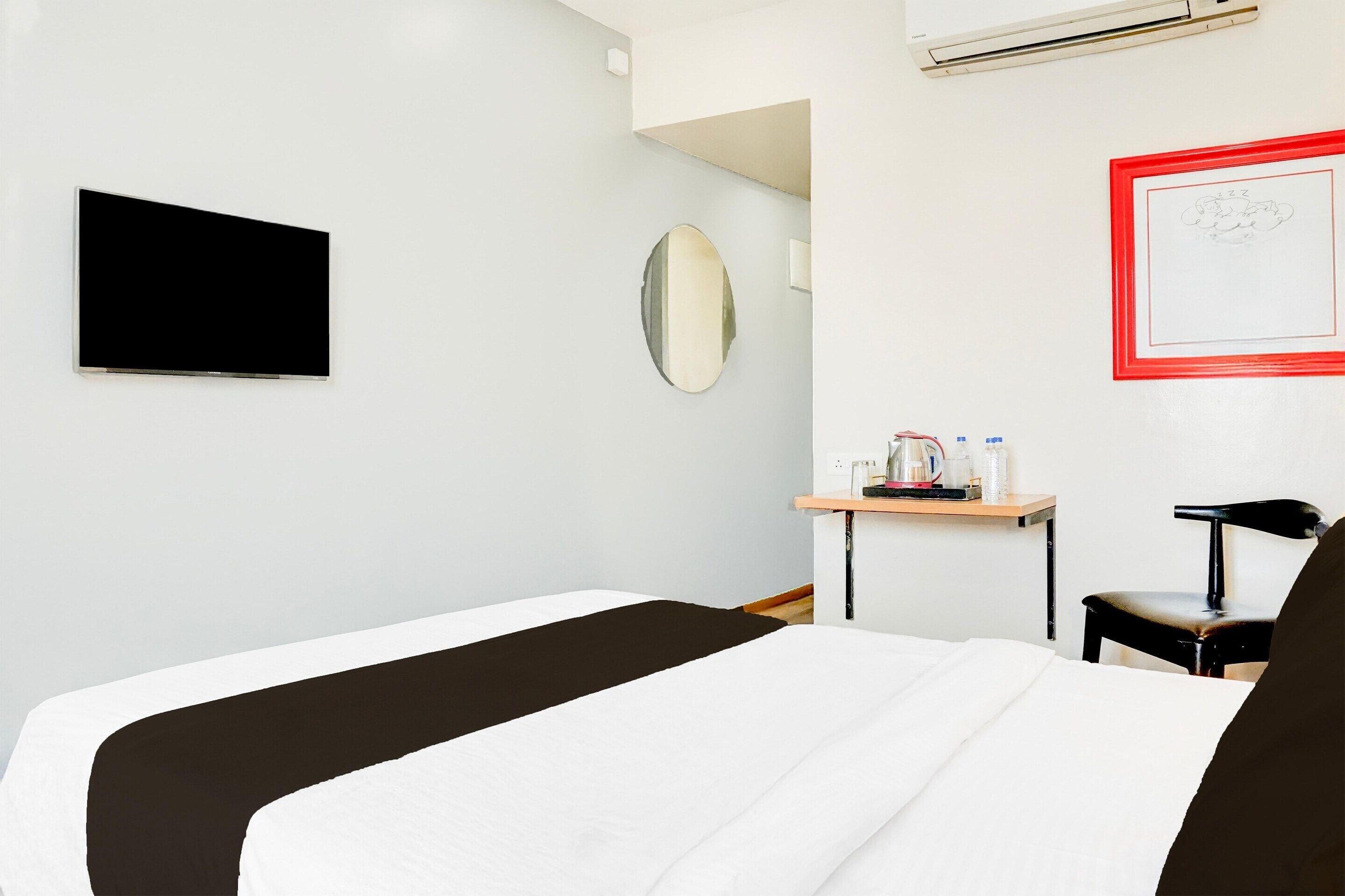 A hotel room with a bed, a desk with a chair, a TV, a mirror, and a framed picture on the wall.