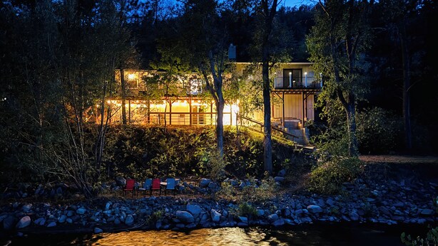 Une maison moderne dotée d’une grande fenêtre donnant sur une rivière, entourée d’arbres et de rochers.