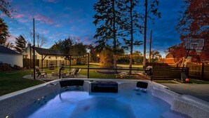A hot tub with a wooden deck and chairs, surrounded by a backyard with a basketball hoop and a wooden fence.