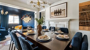 A dining area with a long table set for six, blue upholstered chairs, a chandelier, and a framed photograph on the wall.