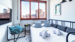 Una habitación de hotel moderna con un ventanal grande, una cama con sábanas blancas, almohadas a rayas, una silla de metal negro y una mesita con un objeto amarillo.
