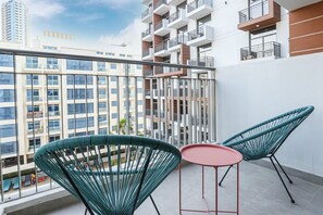 A balcony with two green chairs and a small red table, overlooking a cityscape with modern buildings.