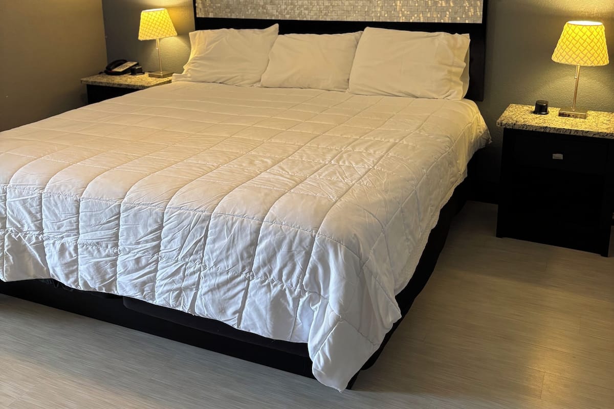 Una cama bien hecha con colcha blanca, dos lámparas de noche, un ventilador de techo y una mesita de noche con un teléfono.