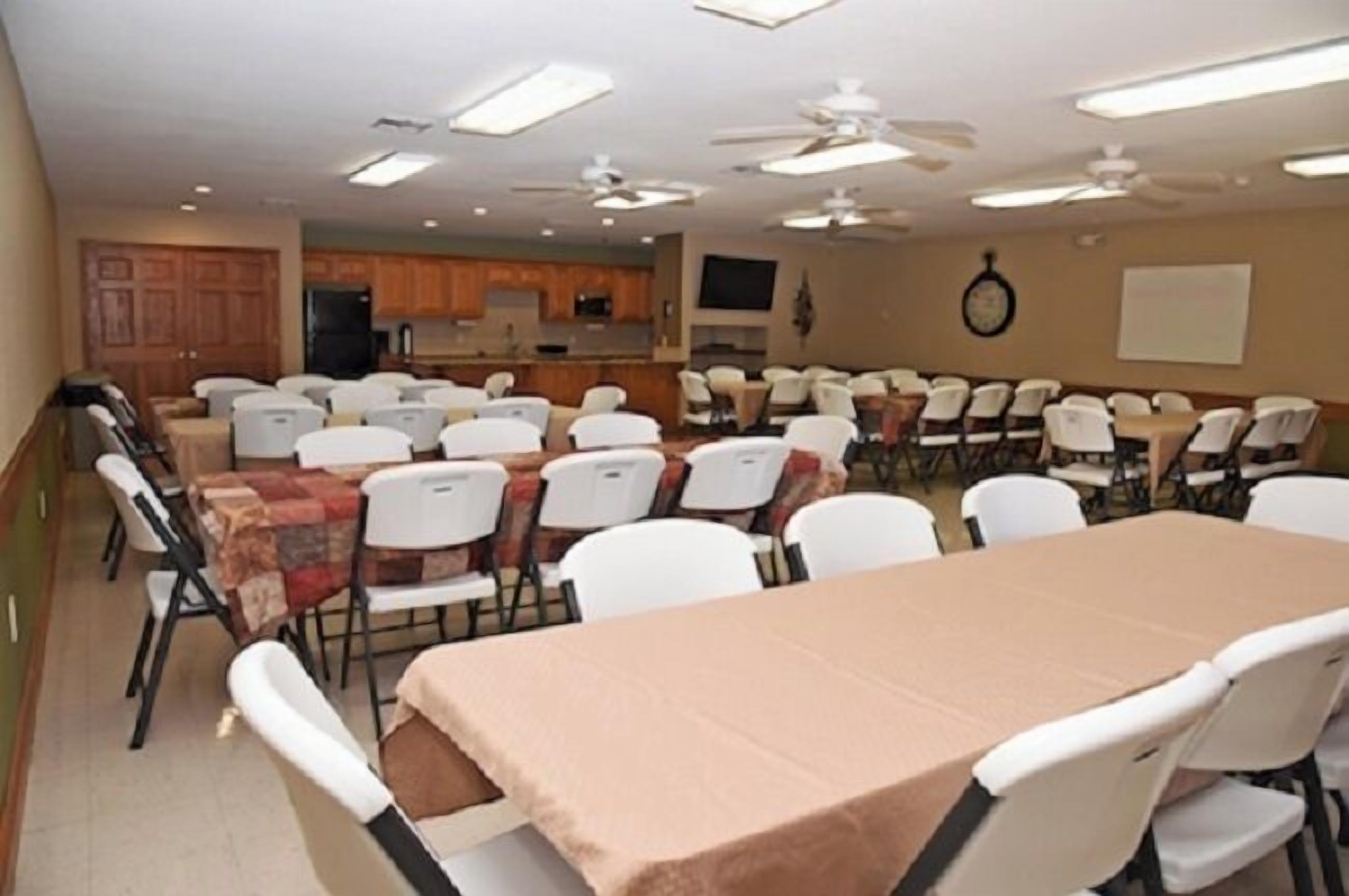 Une salle de conférence équipée de plusieurs tables et chaises, d’un coin cuisine et de ventilateurs de plafond.