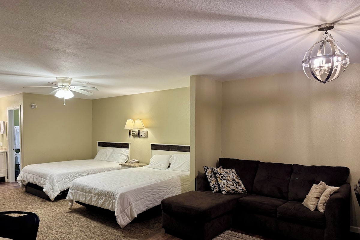 Una habitación de hotel con dos camas, un sofá marrón oscuro, una mesita y un ventilador de techo.