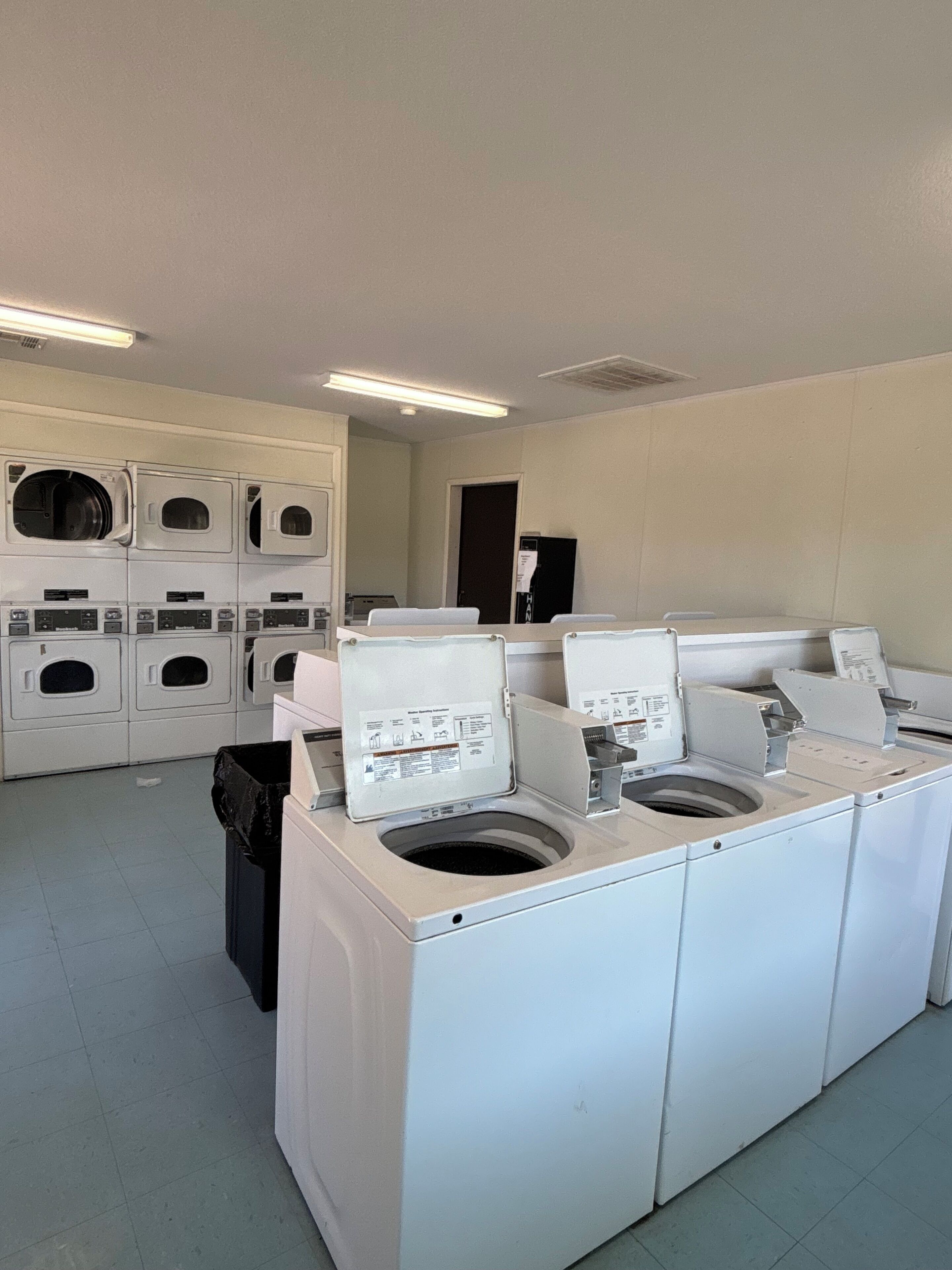 A laundry room with multiple washing machines and dryers, a trash bin, and a door in the background.