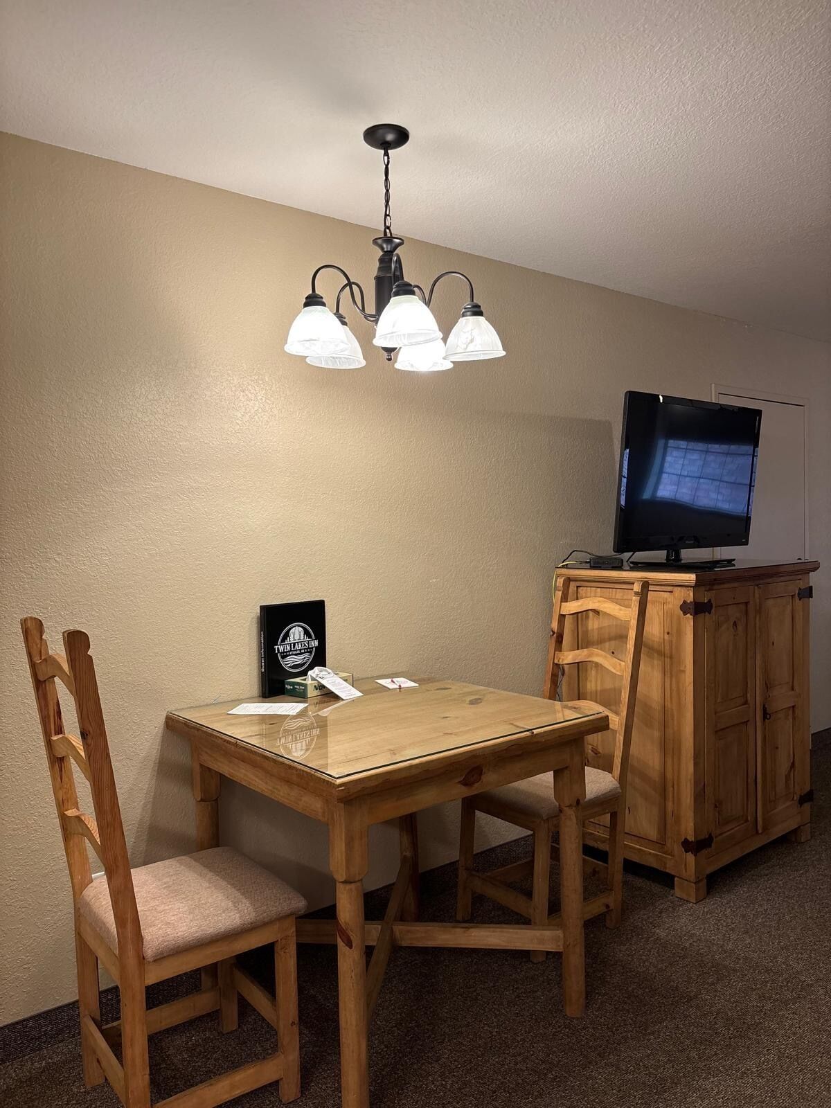 A dining area with a wooden table and chairs, a chandelier, a television on a wooden cabinet, and a framed certificate on the table.