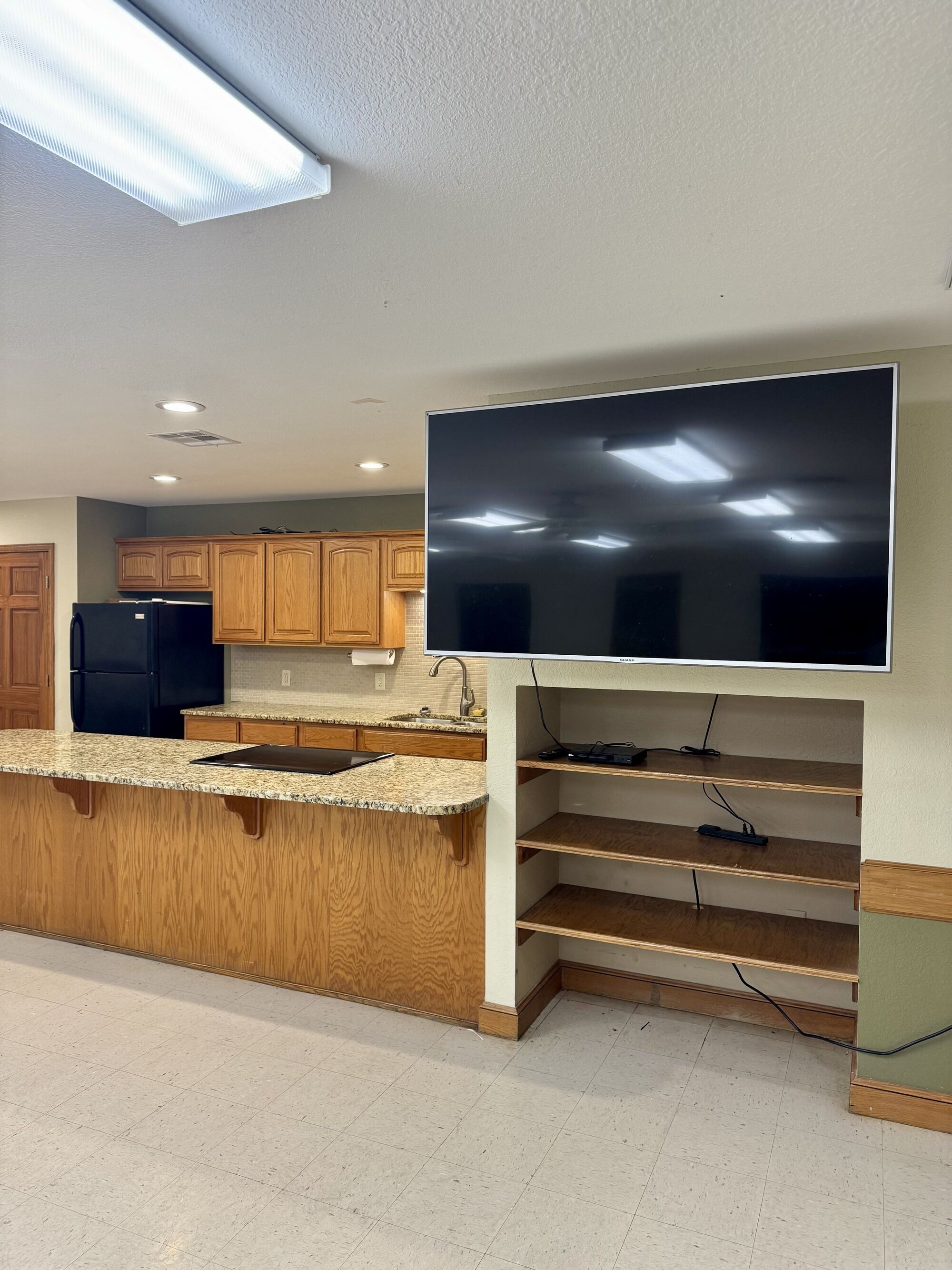 A kitchen with wooden cabinets, a black refrigerator, a granite countertop, and a large flat-screen TV mounted on the wall.