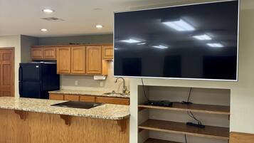 A kitchen with wooden cabinets, a black refrigerator, a granite countertop, and a large flat-screen TV mounted on the wall.