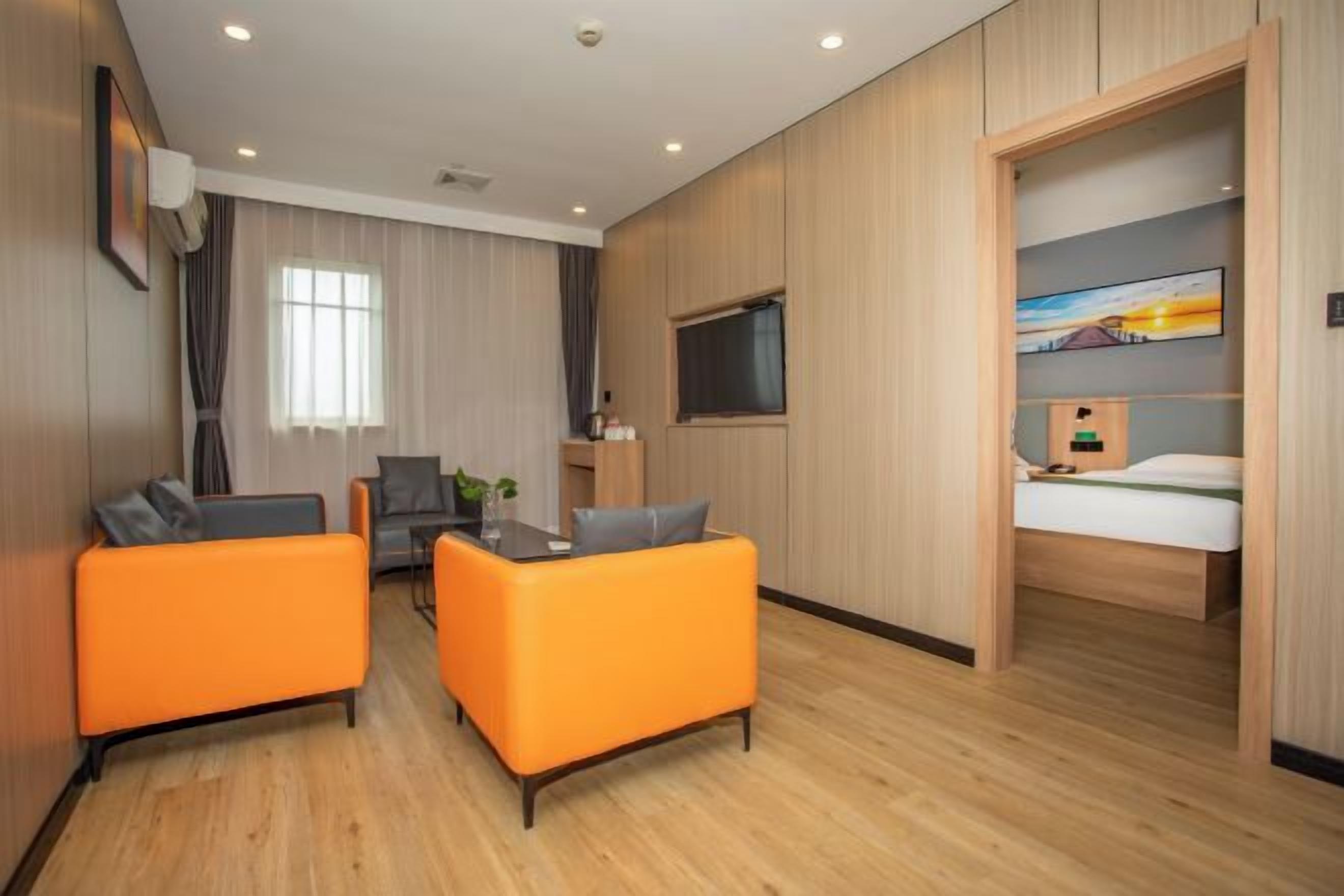 Una habitación de hotel moderna con suelo de madera, sofás naranjas, una mesa de comedor, un televisor y una cama con un cuadro encima.