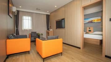 Una habitación de hotel moderna con suelo de madera, sofás naranjas, una mesa de comedor, un televisor y una cama con un cuadro encima.