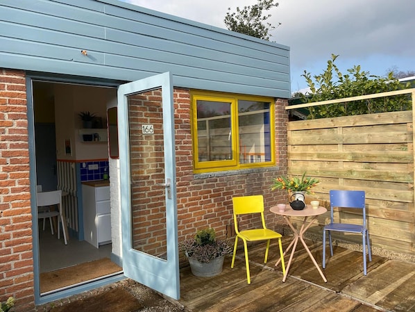 A brick house with a blue exterior, a yellow window, and a door leading to a kitchen. There are two chairs and a small table outside.