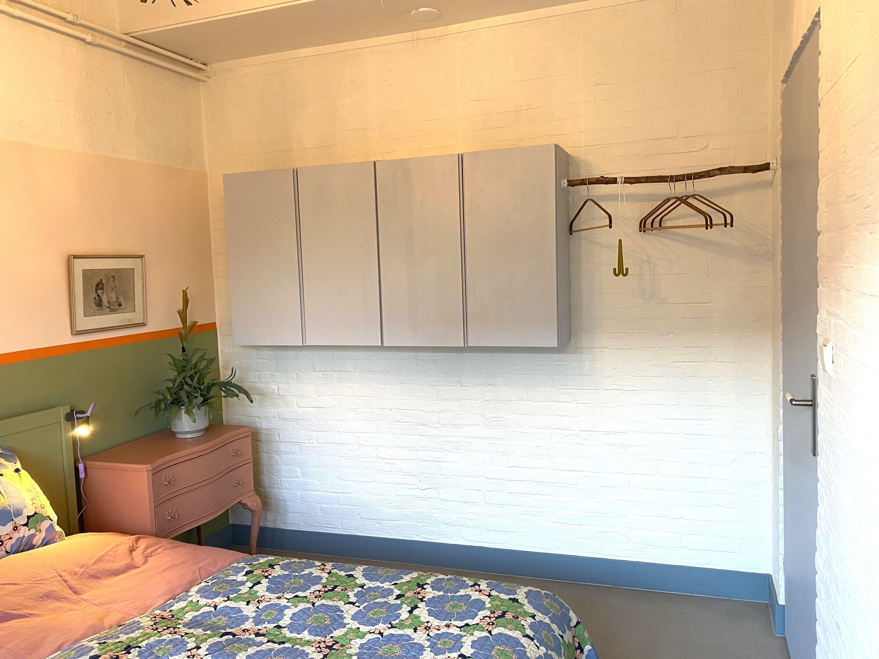 Ein Schlafzimmer mit Bett, Kommode, Kleiderschrank, einer Pflanze und einem Kleiderständer.