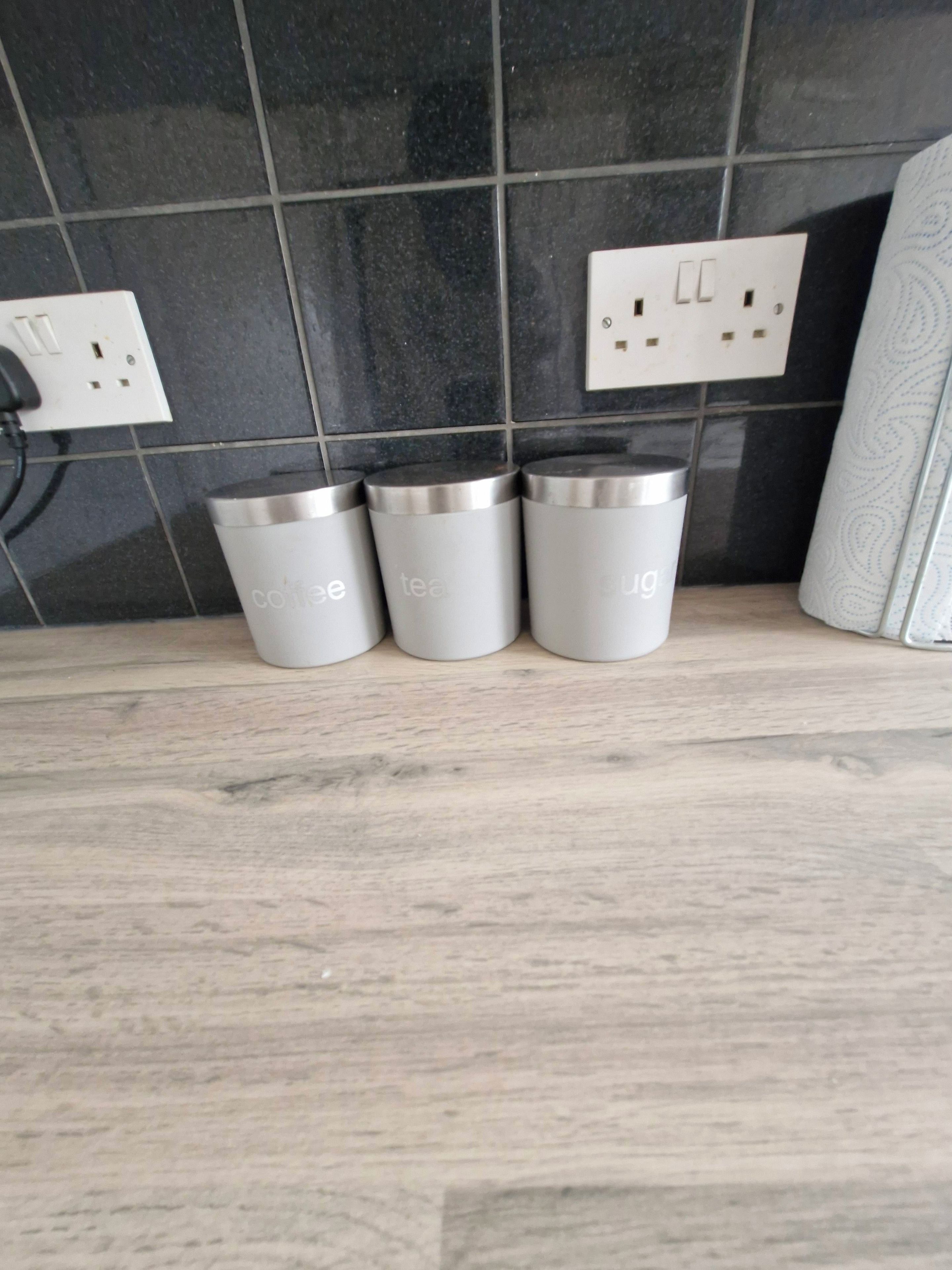 Three stainless steel containers labeled 'coffee,' 'tea,' and 'sugar' on a kitchen counter with a tiled backsplash and electrical outlets.