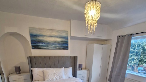 Un dormitorio con una cama grande, mesitas de noche, un armario, un cuadro en la pared y una ventana con cortinas.