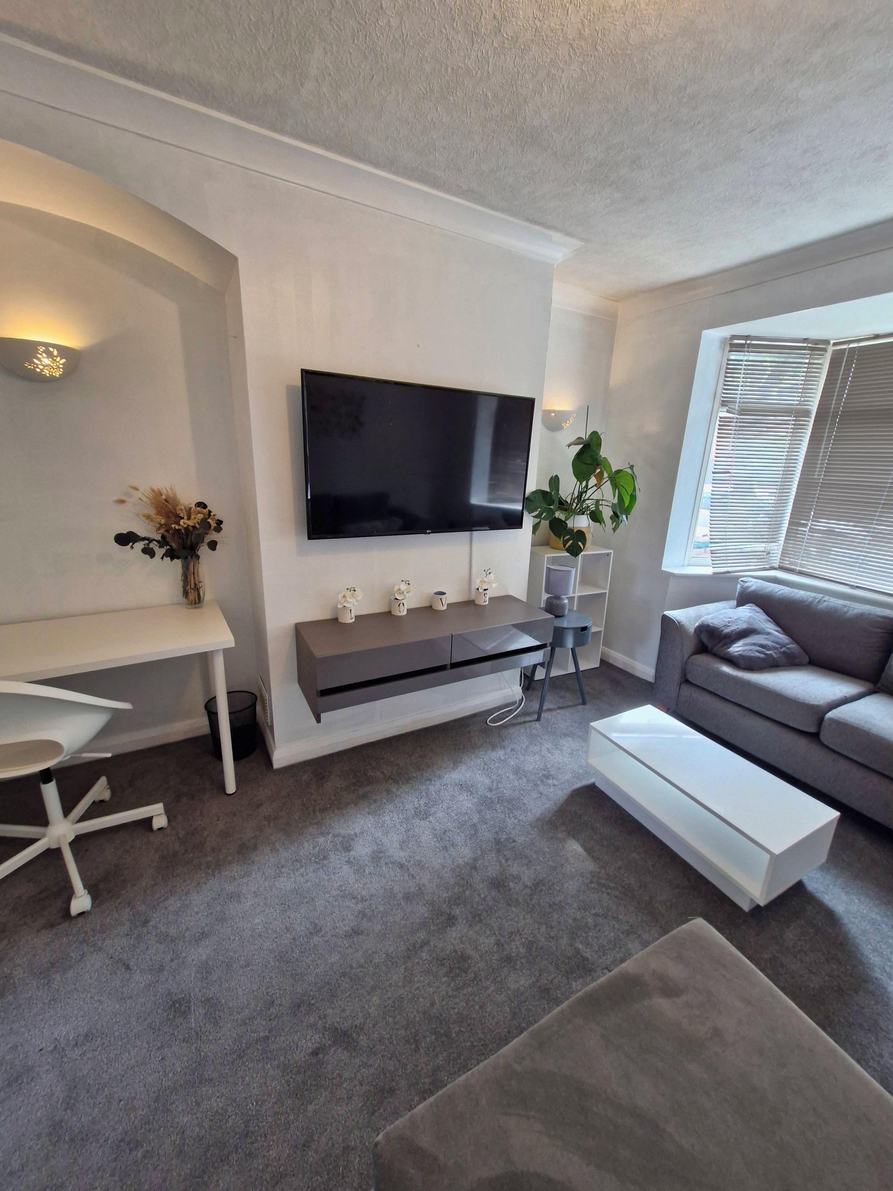 A modern living room with a flat-screen TV mounted on the wall, a grey sofa, a white coffee table, and a small side table with a plant.