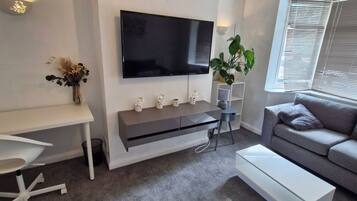 A modern living room with a flat-screen TV mounted on the wall, a grey sofa, a white coffee table, and a small side table with a plant.