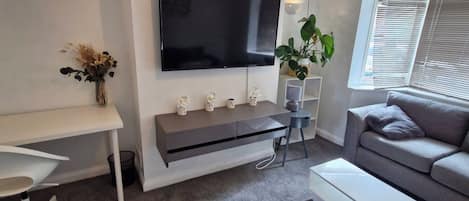 A modern living room with a flat-screen TV mounted on the wall, a grey sofa, a white coffee table, and a small side table with a plant.