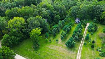 An aerial view of a rural property with a single house surrounded by dense forest, a clear path leading to the house, and a dirt road adjacent to the property.