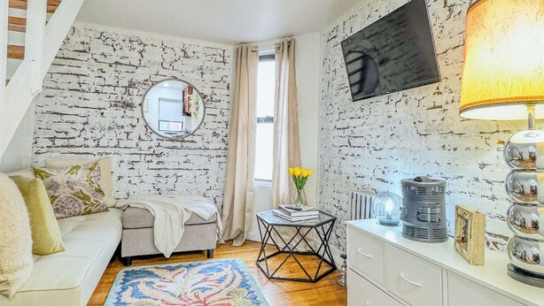 A cozy living room with a white sofa, a patterned rug, a small table with books and flowers, a TV on a brick-patterned wall, and a staircase. - 183 Orchard St (New York)