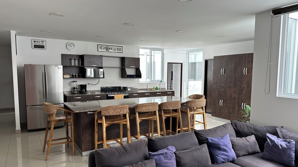A modern kitchen with a central island, bar stools, and a grey sofa in the living area. - Family House 6 Private Rooms, Antiguo Cuscatlán (Antiguo Cuscatlan, Zona Uca)