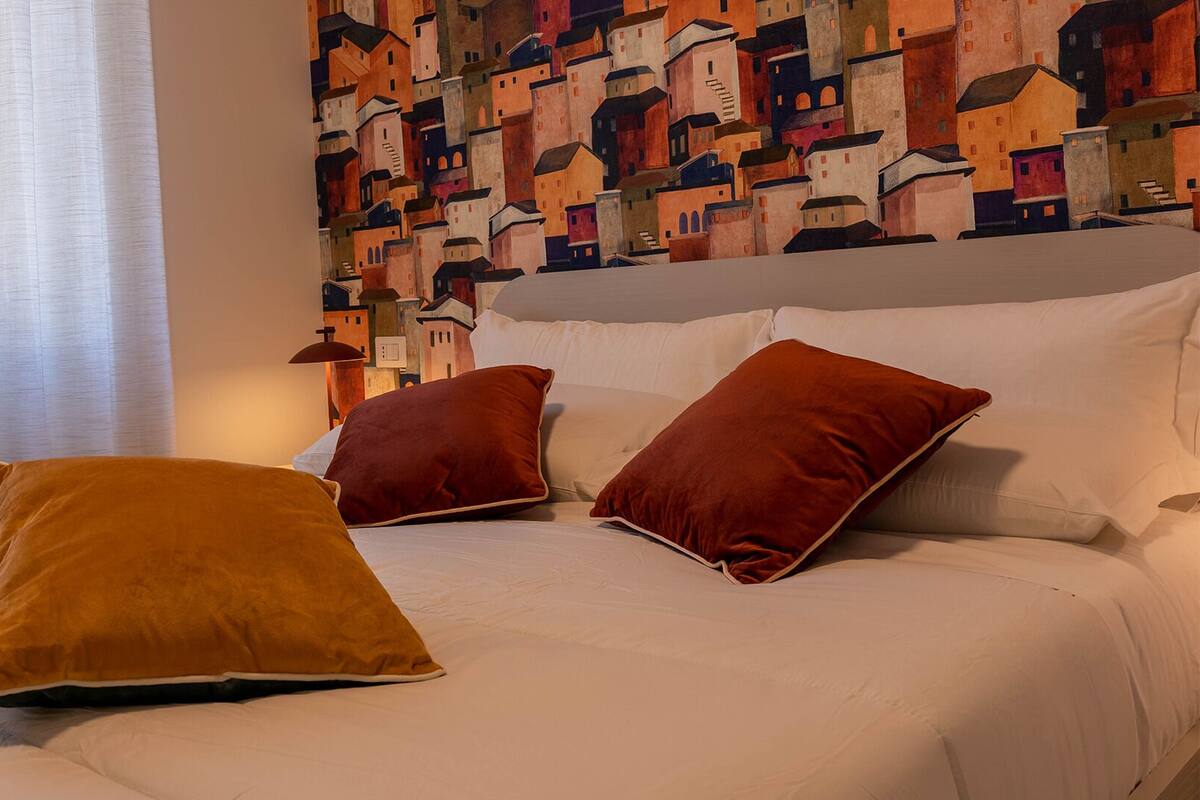 Una cama con ropa blanca y tres almohadas rojas, una ventana con cortinas de gasa y una pared con un mural abstracto de colores.