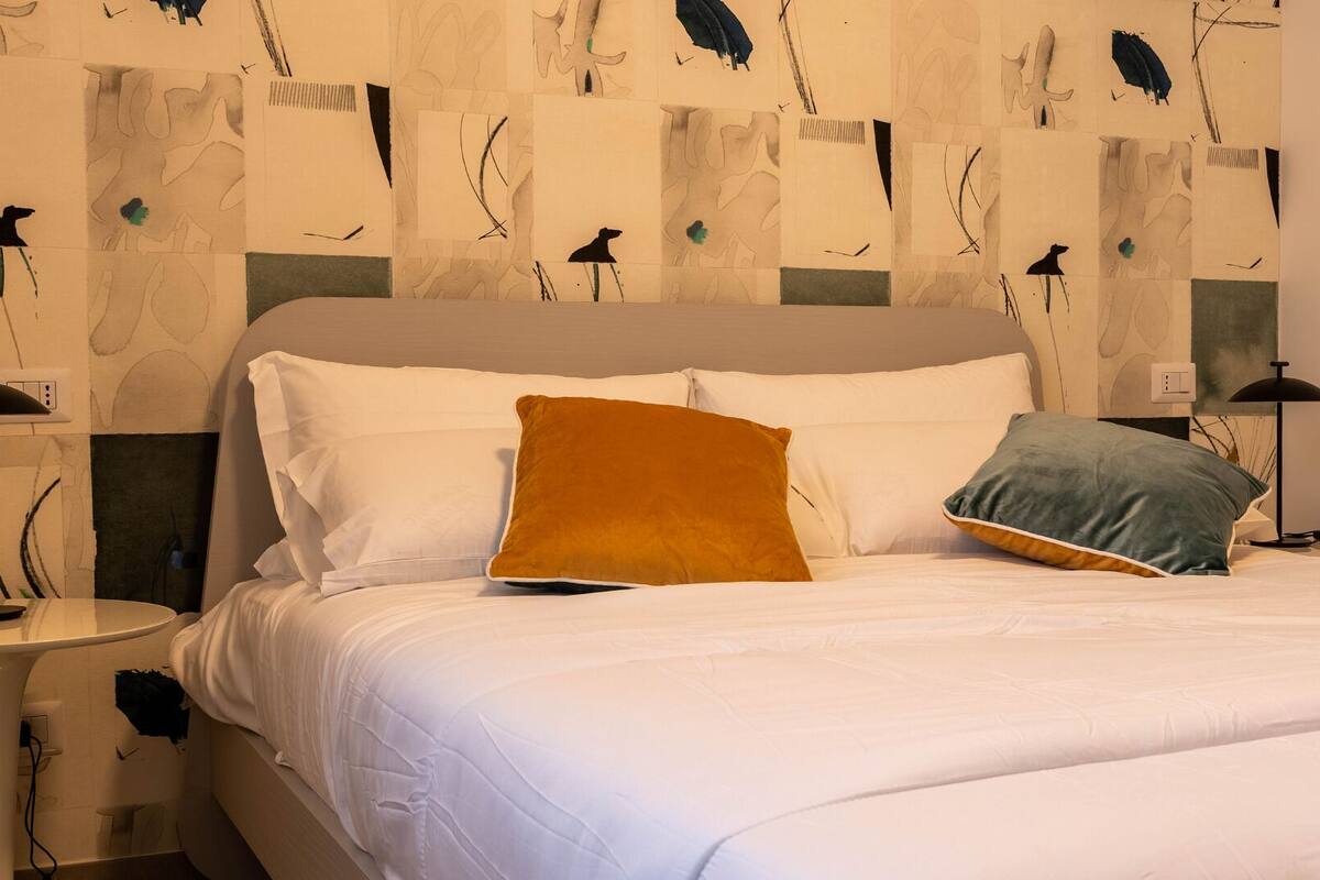 Una cama bien hecha con ropa blanca y dos almohadas, una marrón y otra verde, en una habitación con un moderno cuadro abstracto en la pared y una mesita de noche con una lámpara.