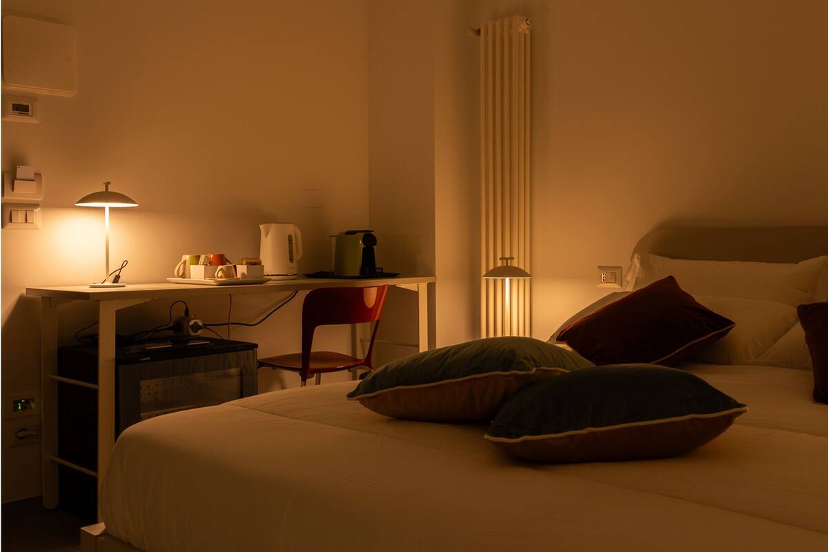 Una habitación de hotel con cama, escritorio, silla, lámpara y radiador.