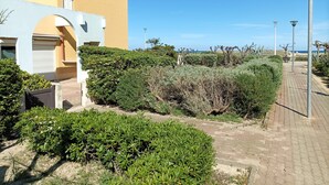 A courtyard with a paved walkway, a building with a yellow facade, and a view of the sea. - Le 108: Ground floor apartment with large terrace on the seafront (Leucate)
