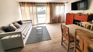 A living room with a sofa, a dining table, and a TV mounted on the wall. - Le 108: Ground floor apartment with large terrace on the seafront (Leucate)