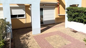 A courtyard with a paved walkway, a white archway, and a closed shuttered window. - Le 108: Ground floor apartment with large terrace on the seafront (Leucate)