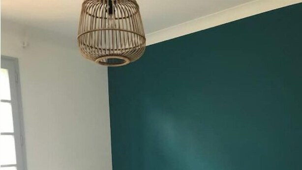 Een netjes opgemaakt bed met witte lakens en een deken met blauw-wit patroon, een muur in teal, een houten vloer en een hanglamp.